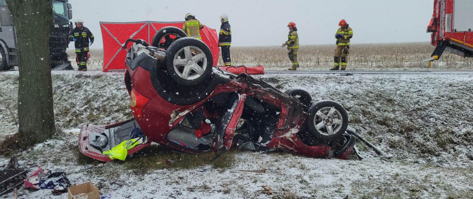 Zdjęcie przedstawia zniszczony samochód po wypadku. Samochód znajduje się w rowie na dachu. Samochód jest czerwony. W tle strażacy biorący udział w działaniach ratowniczych.