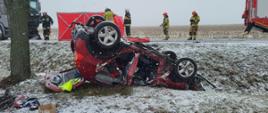 Zdjęcie przedstawia zniszczony samochód po wypadku. Samochód znajduje się w rowie na dachu. Samochód jest czerwony. W tle strażacy biorący udział w działaniach ratowniczych.