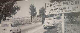 Zdjęcie czarno-białe, samochód mijający tablicę wjazdową do Wrocławia z informacją o zakazie wjazdu niezaszczepionym z powodu epidemii ospy