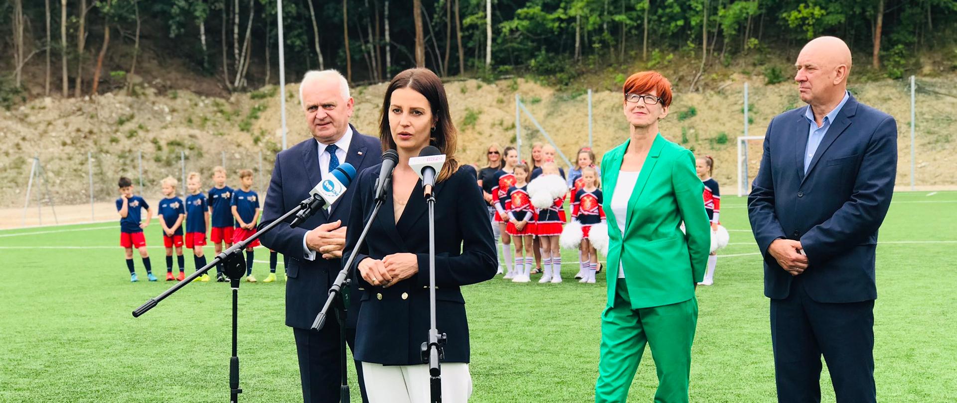 Minister Danuta Dmowska-Andrzejuk stoi przed mikrofonami, w tle pozostali członkowie konferencji, w tym poseł do Parlamentu Europejskiego Elżbieta Rafalska. Za nimi grupa dzieci.
