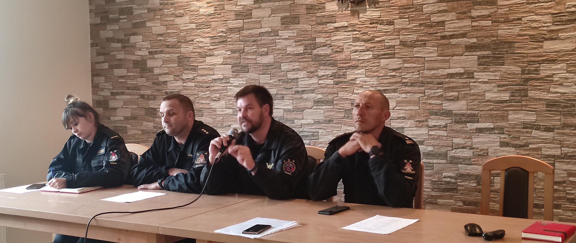 Przy stole siedzą 4 funkcjonariusze, w tle godło na ścianie. Jeden z nich przemawia przez mikrofon.