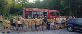 Zdjęcie przedstawia spotkanie strażaków JRG przy KP PSP z Bochni z uczestnikami obozu Młodzieżowych Drużyn Pożarniczych. Na zdjęciu widać czerwony samochód pożarniczy który ma otwarte skrytki. Przy samochodzie stoi strażak i przedstawia sprzęt będący na wyposażeniu pojazdu. Przed pojazdem tyłem stoją dzieci biorące udział w obozie ubrane w piaskowe i czarne koszulki z napisem straż. Po prawej stronie widać część pojazdu osobowego