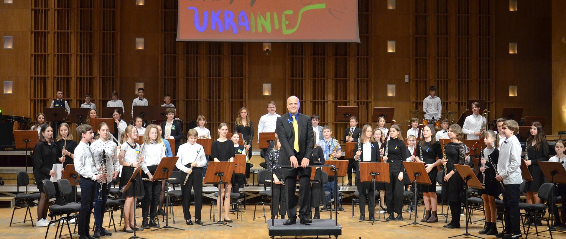 Kolorowe zdjęcie przedstawiające orkiestrę dętą. Na środku sceny, na podeście stoi dyrygent orkiestry a obok niego i za nim stoją uczniowie trzymający swoje instrumenty. Wszyscy wykonawcy ubrani są w czarno- białe stroje. Na scenie znajdują się również pulpity do nut. Ponadto pod sufitem wisi ekran na którym wyświetlony jest napis muzycy Ukrainie.