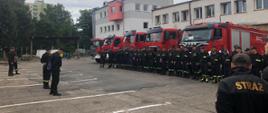 Zdjęcie przedstawia zbiórkę strażaków podczas kompletowania warmińsko-mazurskiej brygady odwodowej w Elblągu. Strażacy ustawieni w szeregu, ubrani w czarne ubranie koszarowe, czerwone samochody strażackie ustawione w jednej linii.
