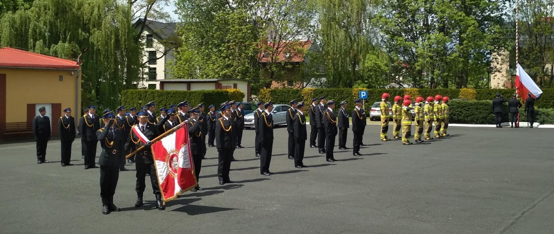 Na zdjęciu strażacy PSP w mundurach wyjściowych, na początku po lewej stronie poczet sztandarowy pochyla sztandar, dalej strażacy w mundurach specjalnych i czerwonych hełmach, na samym końcu poczet flagowy wciąga flagę państwową.