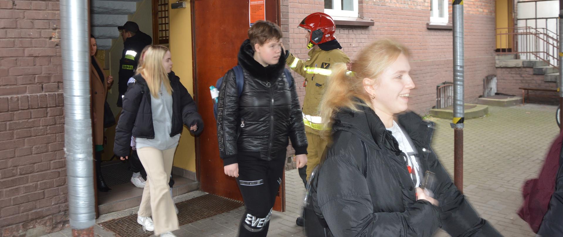 Ćwiczenia w ZSZ. Z budynku szkolnego "B" ewakuują się uczniowie i nauczyciele. Mają założone kurtki i niosą plecaki. Przebieg ewakuacji nadzoruje dwóch strażaków - jeden z nich ma na sobie ubranie specjalne koloru piaskowego i czerwony hełm, drugi - ubranie dowódczo-sztabowe i czapkę. W tle fragment budynku.