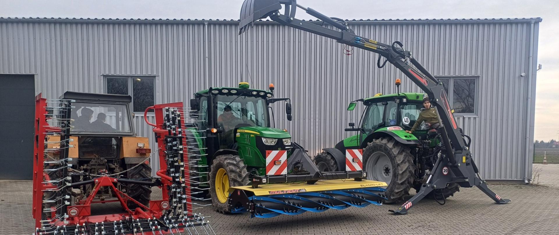 Trzy ciągniki rolnicze z podczepionym osprzętem przed halą z szarej blachy