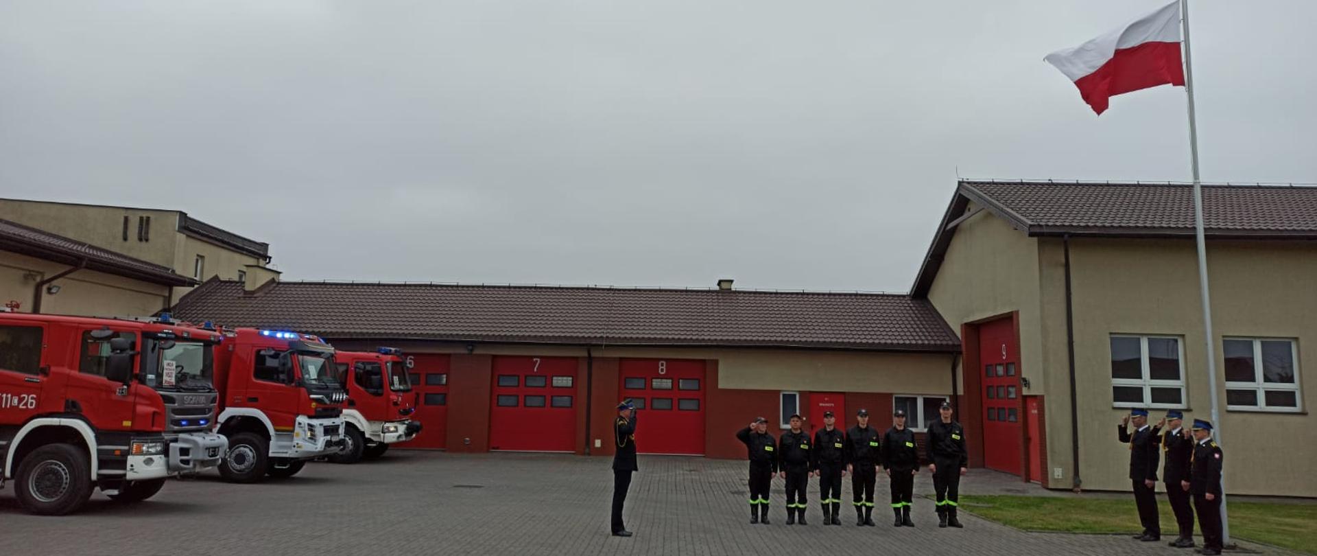 Na zdjęciu widzimy grupę strażaków Państwowej Straży Pożarnej w Rypinie. Znajdują się oni na placu Jednostki Ratowniczo-Gaśniczej. Trzech funkcjonariuszy po wciągnięciu flagi państwowej na maszt oddaje honor. Na przeciwko nich stoi Zastępca Komendanta Państwowej Straży Pożarnej, który wraz z dowódcą pododdziału także oddaje honor. Po lewej stronie znajdują się samochody gaśnicze, a w tle widzimy garaże.