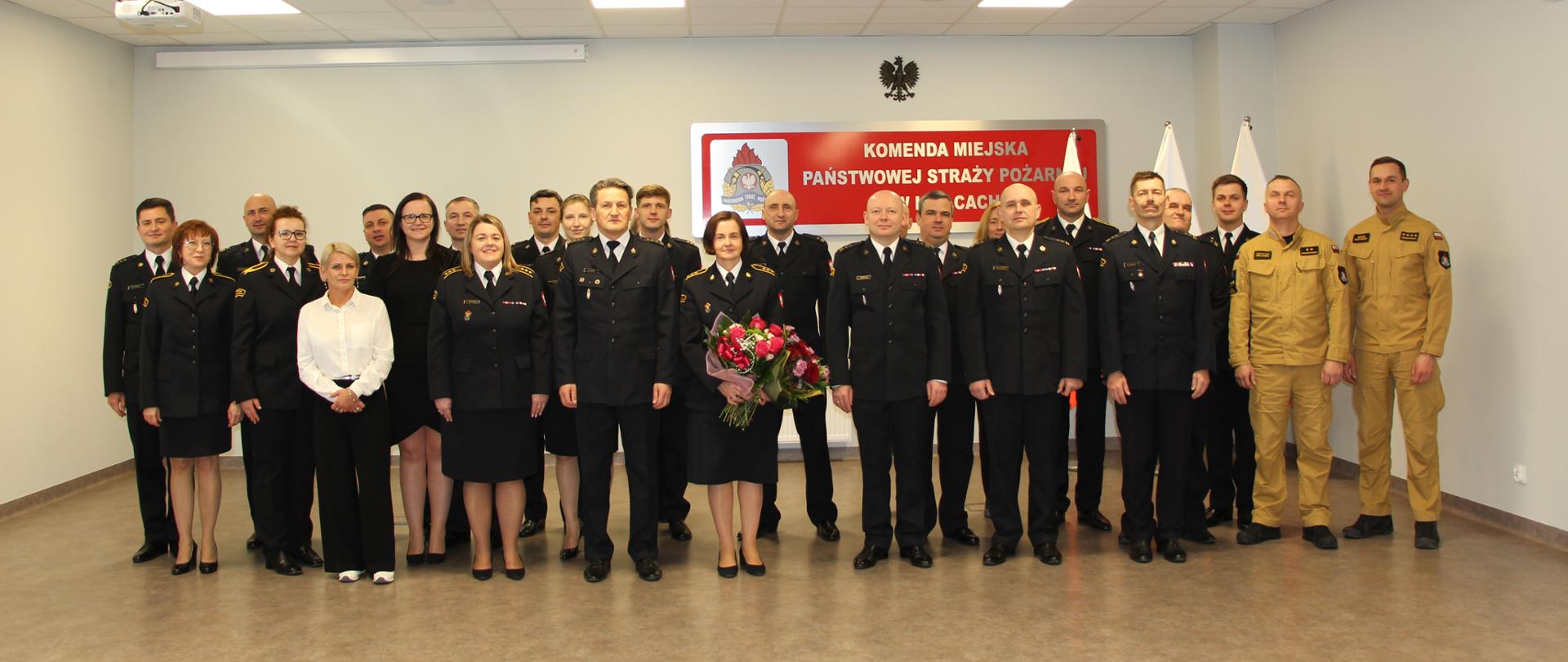Fotografia przedstawia funkcjonariuszy i pracowników cywilnych ustawionych do pamiątkowego zdjęcia. W pierwszym rzędzie stoi kadra kierownicza tutejszej komendy oraz opuszczająca szeregi funkcjonariuszka.