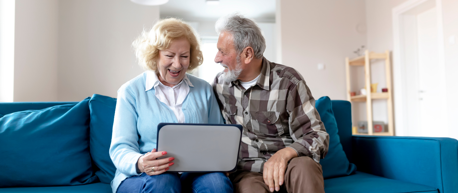 Uśmiechnięci seniorzy - kobieta i mężczyzna, siedzą na kanapie. Kobieta trzyma w ręku laptop, mężczyzna patrzy na kobietę.