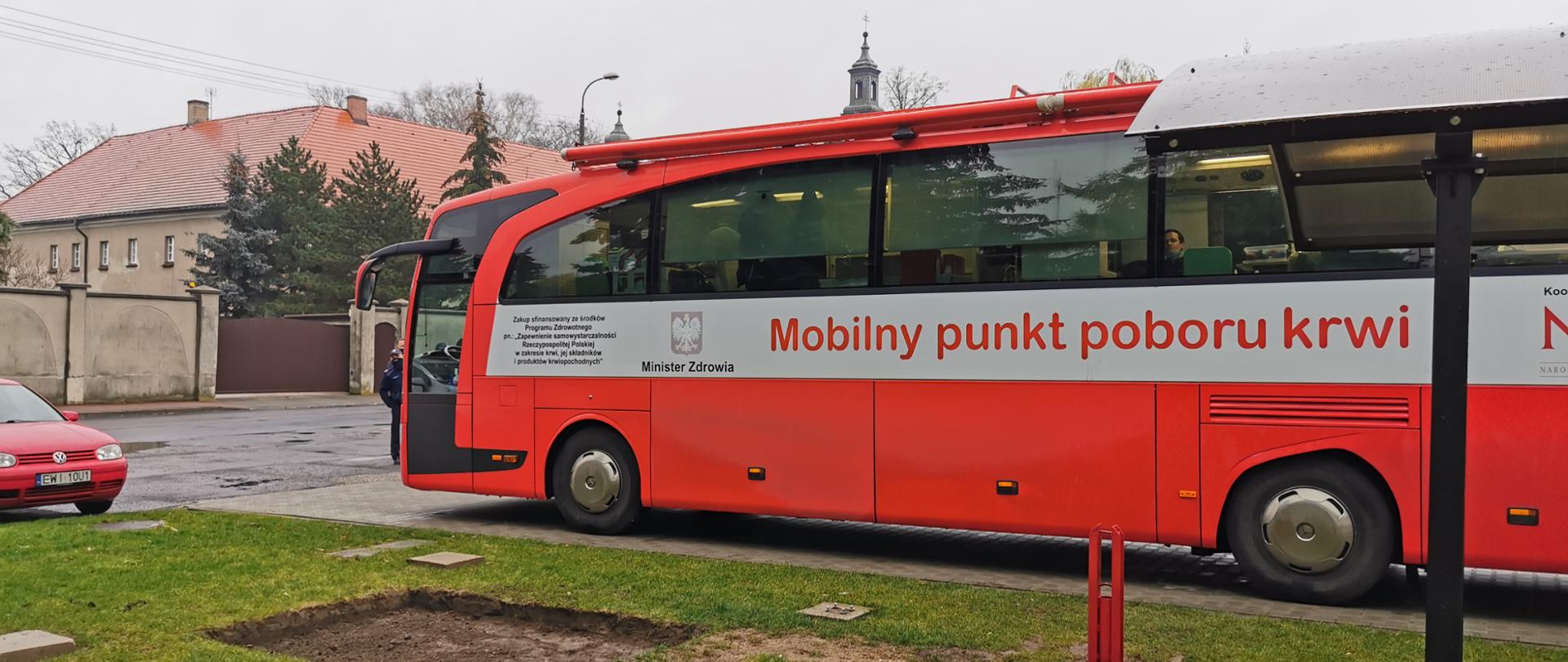 Zdjęcie przedstawia czerwony autobus do mobilnego poboru krwi, w tle klasztor ojców franciszkanów w Wieluniu z wieżyczką kościoła