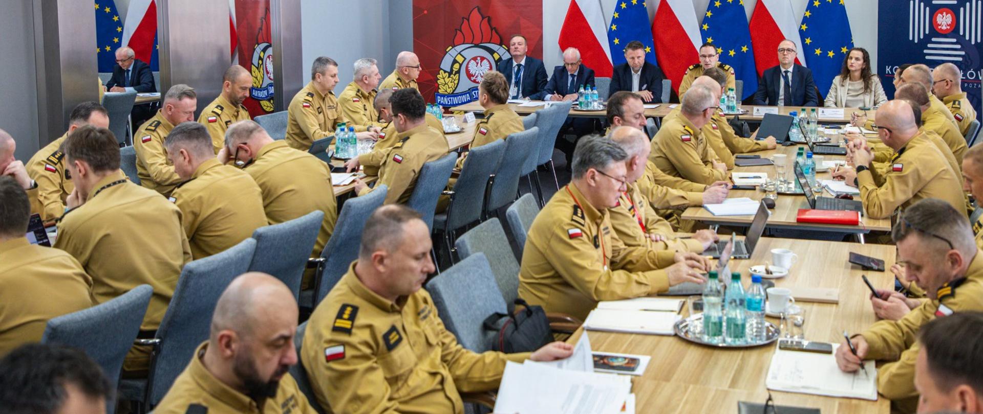 Szerokie ujęcie sali konferencyjnej w Komendzie Głównej PSP; liczni funkcjonariusze siedzą przy stołach i uczestniczą w naradzie.
