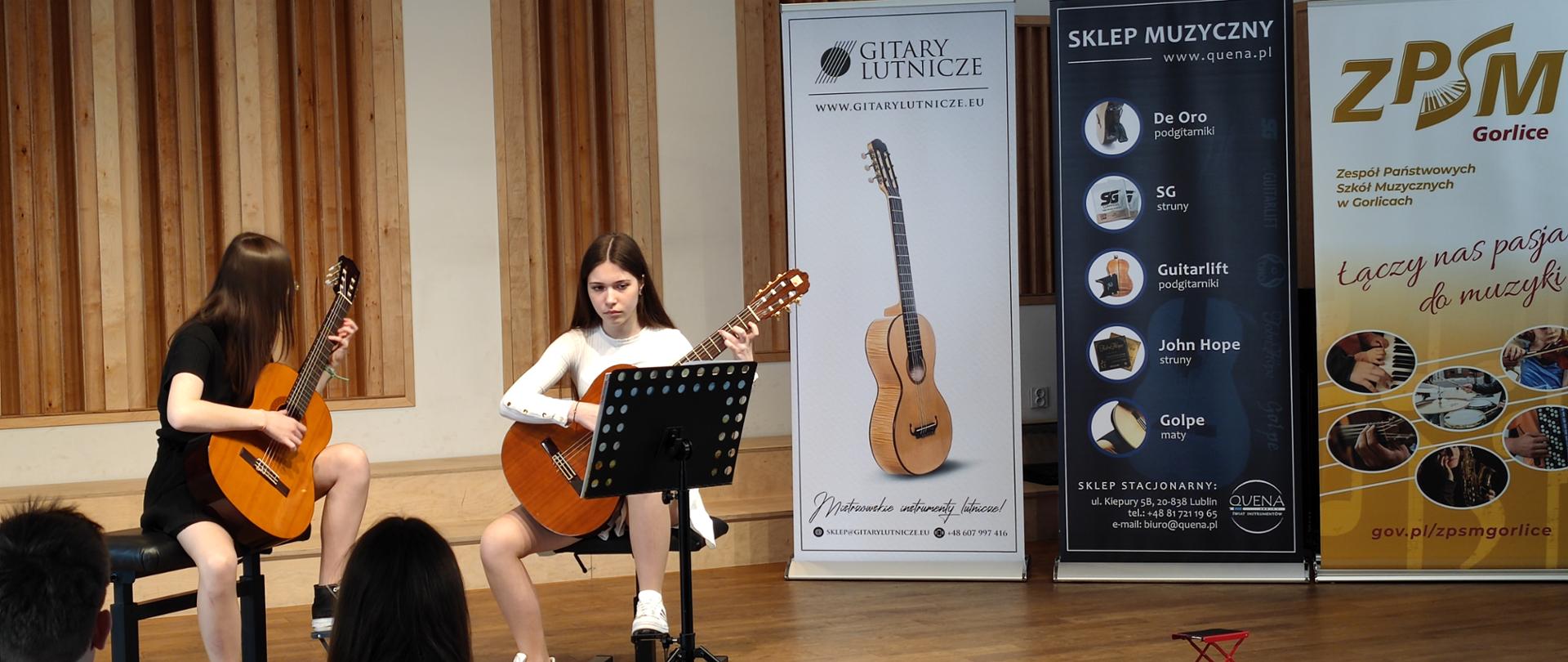 Z lewej strony ubrana na czarno grająca na gitarze dziewczynka z rozpuszczonymi ciemnymi włosami, pośrodku dziewczynka ubrana w białą bluzkę również grające na gitarze. Po prawej stronie stoją banery reklamowe