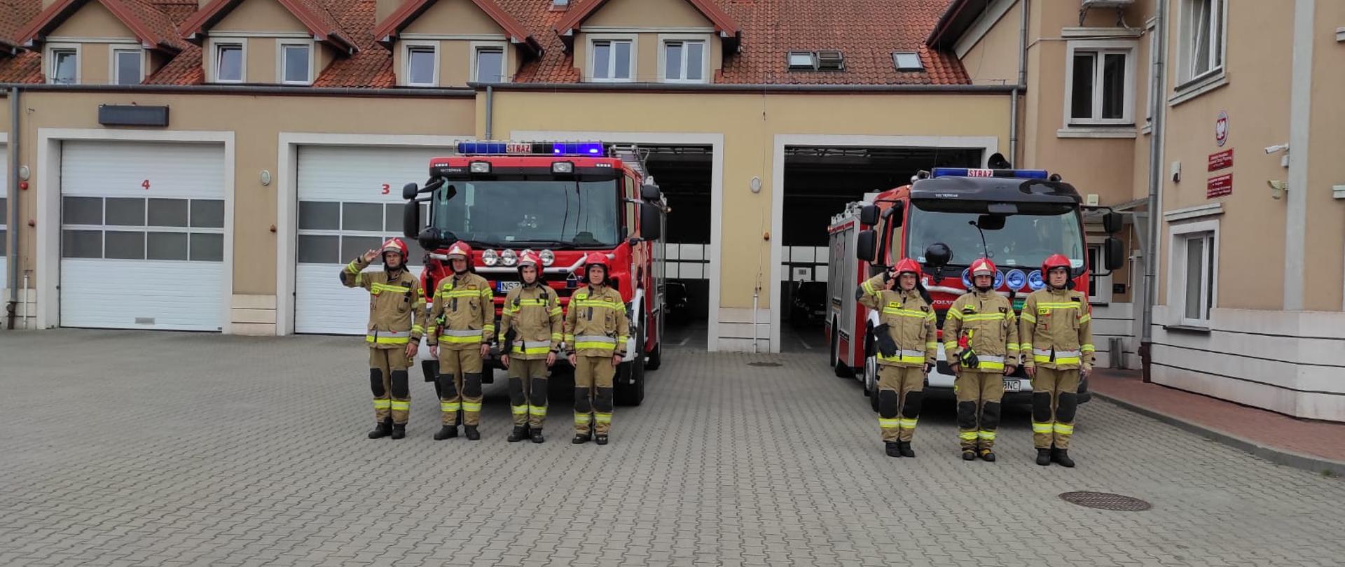 Zdjęcie przedstawia strażaków oddających honor z okazji rocznicy Powstania Warszawskiego na tle samochodów pożarniczych oraz budynku Komendy Powiatowej PSP w Szczytnie