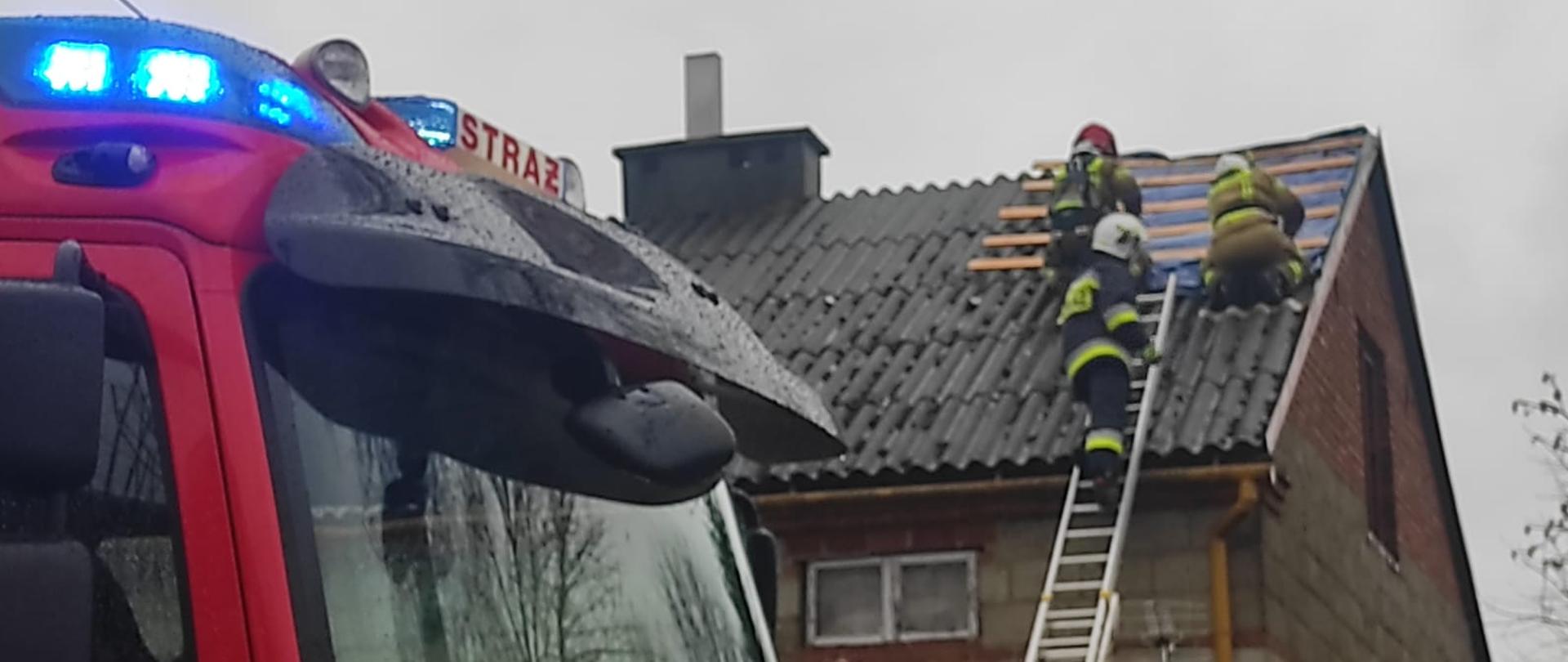 Kolorowa fotografia wykonana w pochmurny dzień, przedstawia na pierwszym planie część kabiny samochodu strażackiego z włączonym niebieskim światłem na dachu. W tle widać szary budynek i trzech strażaków w ubraniach bojowych, zabezpieczających niebieską plandeką uszkodzony dach. O budynek oparta drabina strażacka.