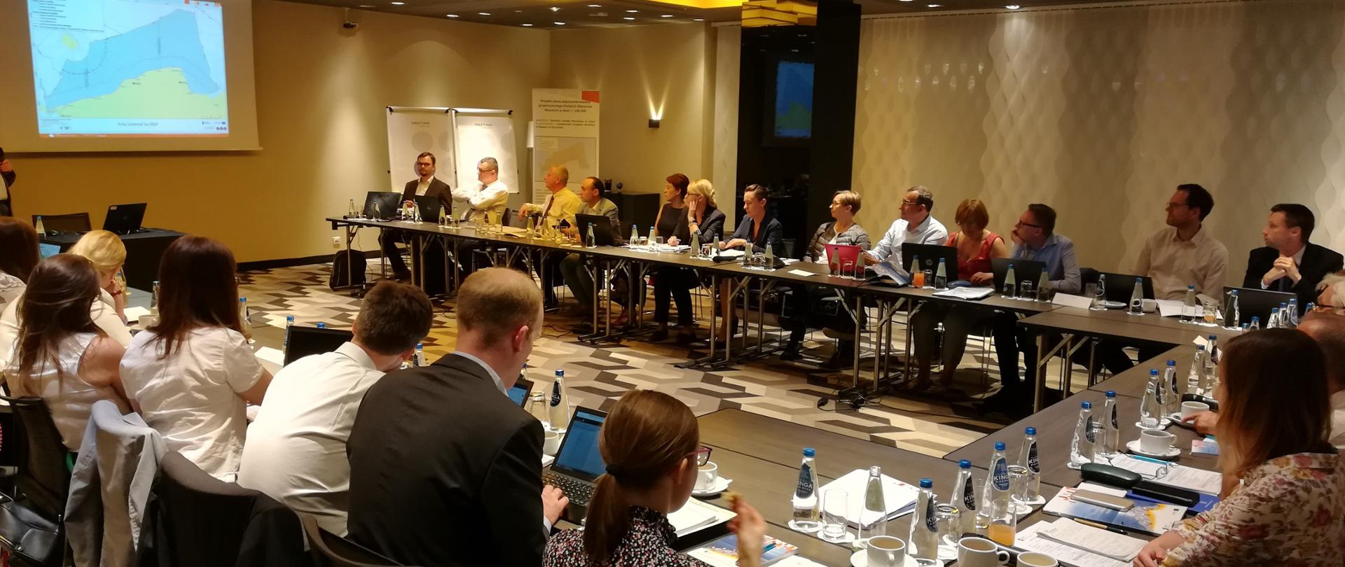 
Uczestnicy spotkania siedzą przy stole, w tle widać prezentację multimedialną.