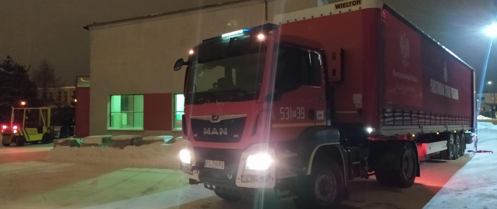 Na zdjęciu widać pojazd ciężarowy z naczepą transportową. Pojazd w kolorze czerwonym, z białymi elementami zderzaka oraz sygnalizacją ostrzegawczą na dachu. Na boku naczepy widnieje napis Państwa Straż Pożarna oraz logo tej formacji mundurowej. W tle widać budynek magazynowy w kolorze biało czerwonym znajdujący się na terenie Jednostki Ratowniczo-Gaśniczej nr 1 w Kaliszu. Obok magazynu widać również wózek widłowy wykorzystywany do rozładunku pojazdu.