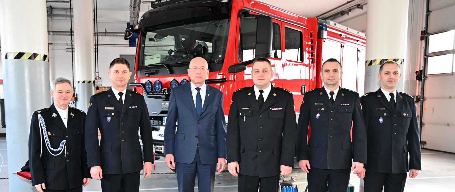 Strażacy na tle samochodu pożarniczego za nimi z tyłu pomarańczowa mata ze sprzętem