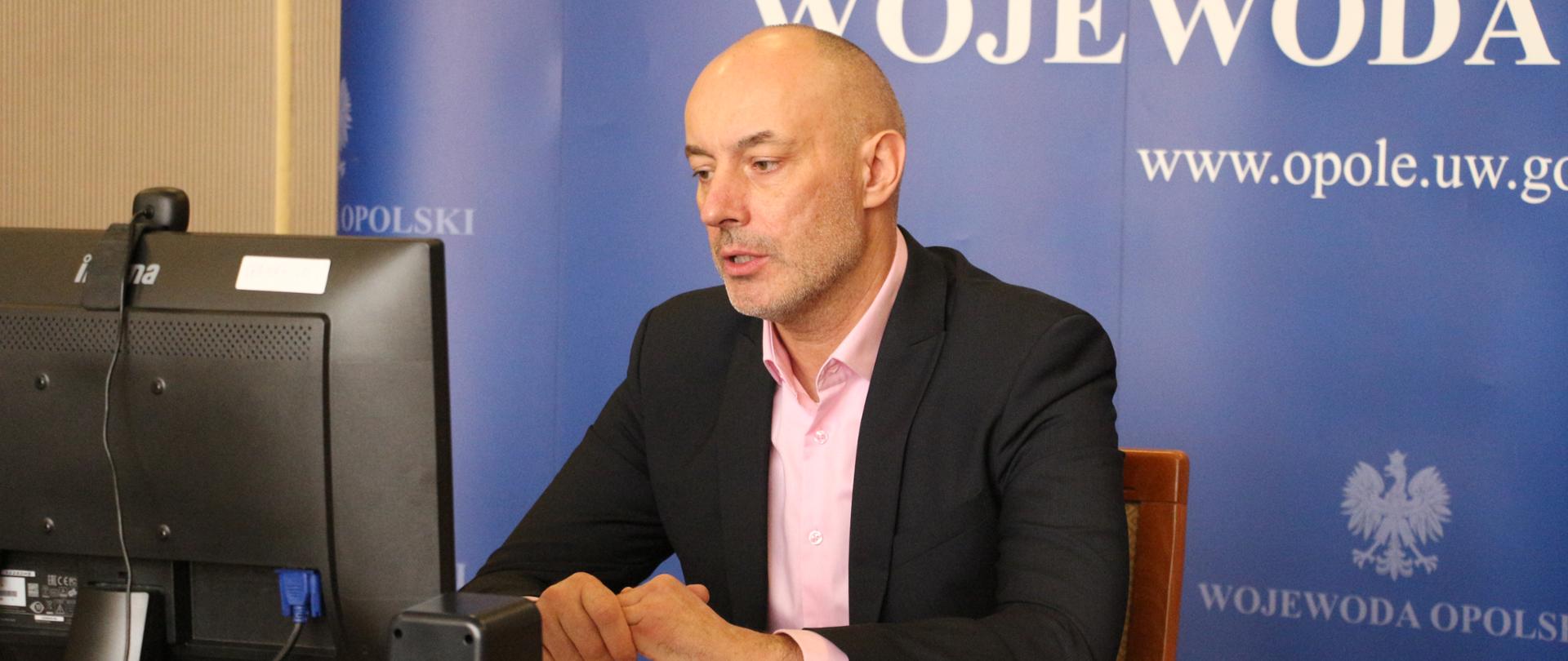 Wojewoda opolski podczas wideokonferencji z mediami. Wojewoda siedzi przy stole, na którym umieszczono duży monitor komputerowy. Wojewoda spogląda na niego, mówi do słuchaczy. Ma splecione dłonie. Za wojewodą ustawiona granatowa ścianka z napisem Wojewoda Opolski oraz godłem.