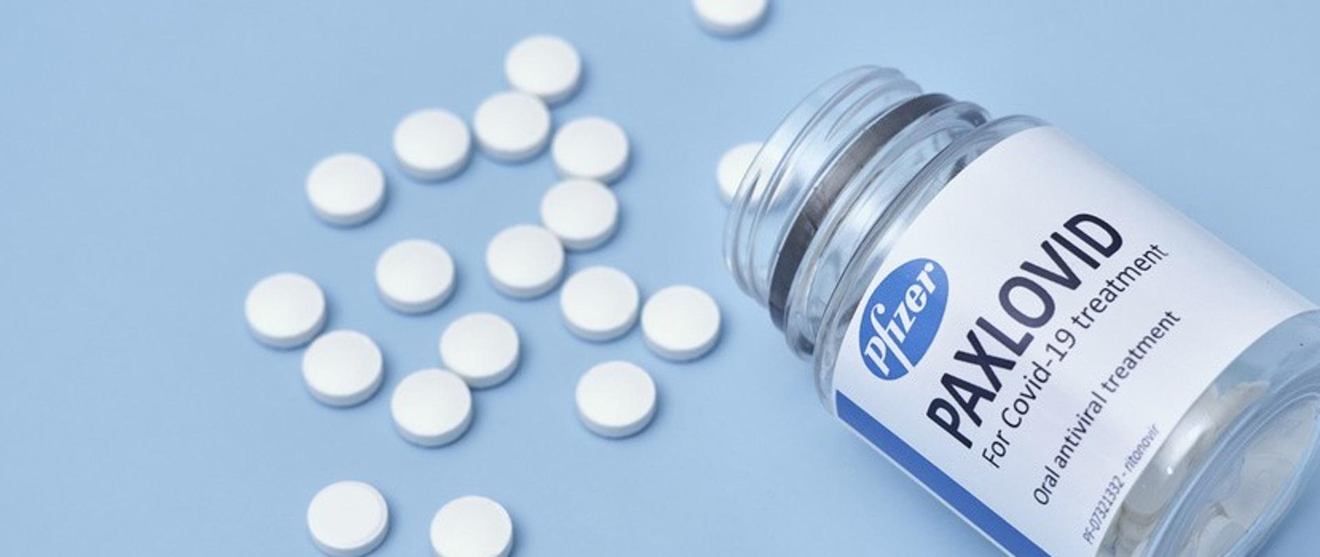 rozsypane białe, okrągłe tabletki oraz słoiczek z napisem paxlovid na jasnoniebieskim tle