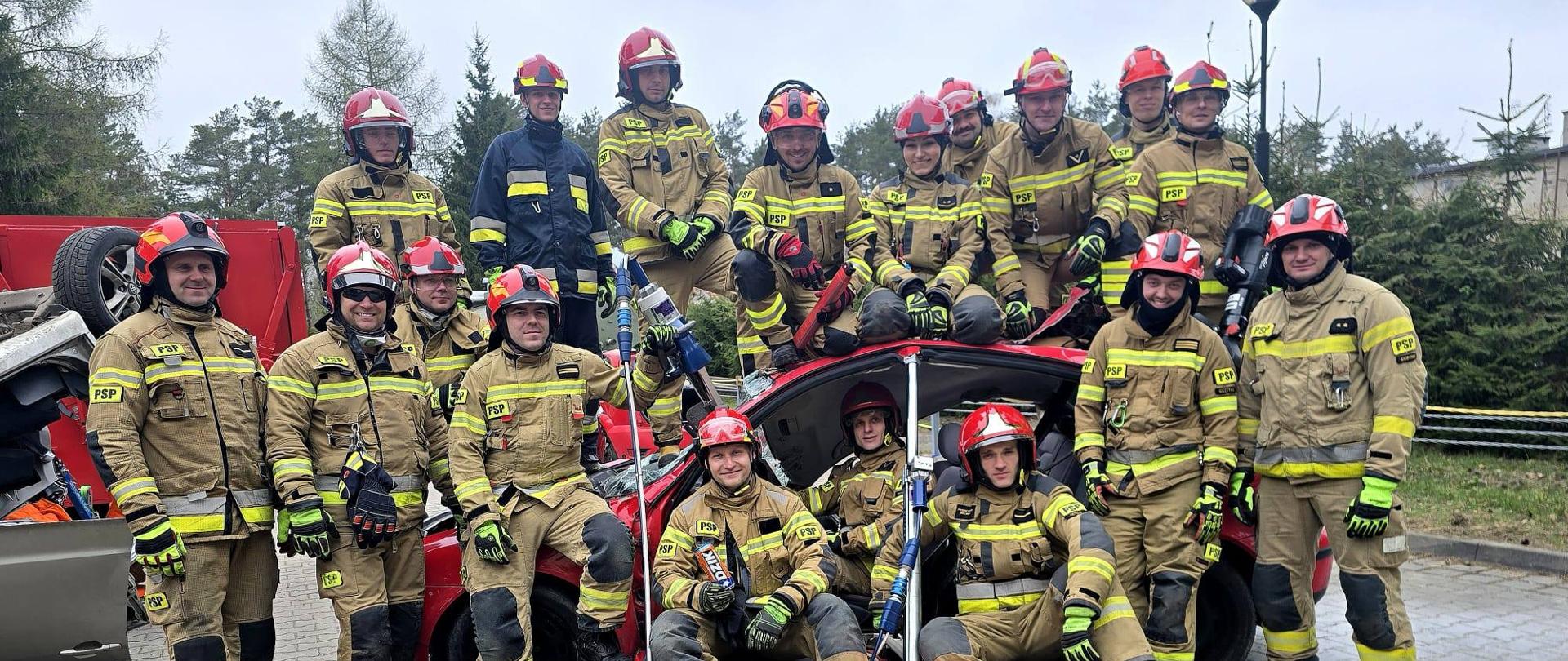 Grupa kilkunastu strażaków w mundurach bojowych i czerwonych hełmach ze sprzętem do ratownictwa technicznego. Pozują do zdjęcia. Część z nich siedzi na wraku samochodu osobowego.