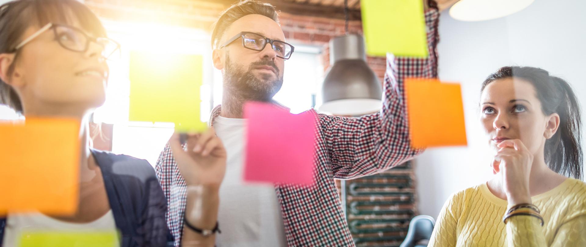 mężczyzna i dwie kobiety stoją przed tablicą na której na kolorowych kartkach zapisują pomysły
