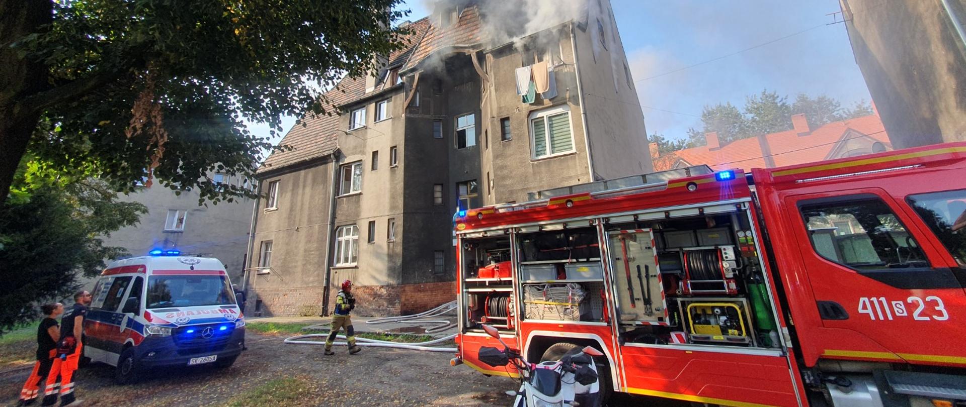 Na zdjęciu widać akcję ratowniczo-gaśniczą prowadzoną przy wielorodzinnym budynku mieszkalnym, z którego dachu wydobywa się gęsty dym. Na miejscu obecne są zastępy straży pożarnej z rozwiniętym sprzętem gaśniczym oraz karetka pogotowia ratunkowego z ratownikami medycznymi. Przed budynkiem stoi wóz strażacki z otwartymi skrytkami, z widocznym sprzętem technicznym i wężami pożarniczymi. Działania prowadzone są w ciągu dnia, a sytuacja wskazuje na intensywny pożar poddasza lub dachu budynku.