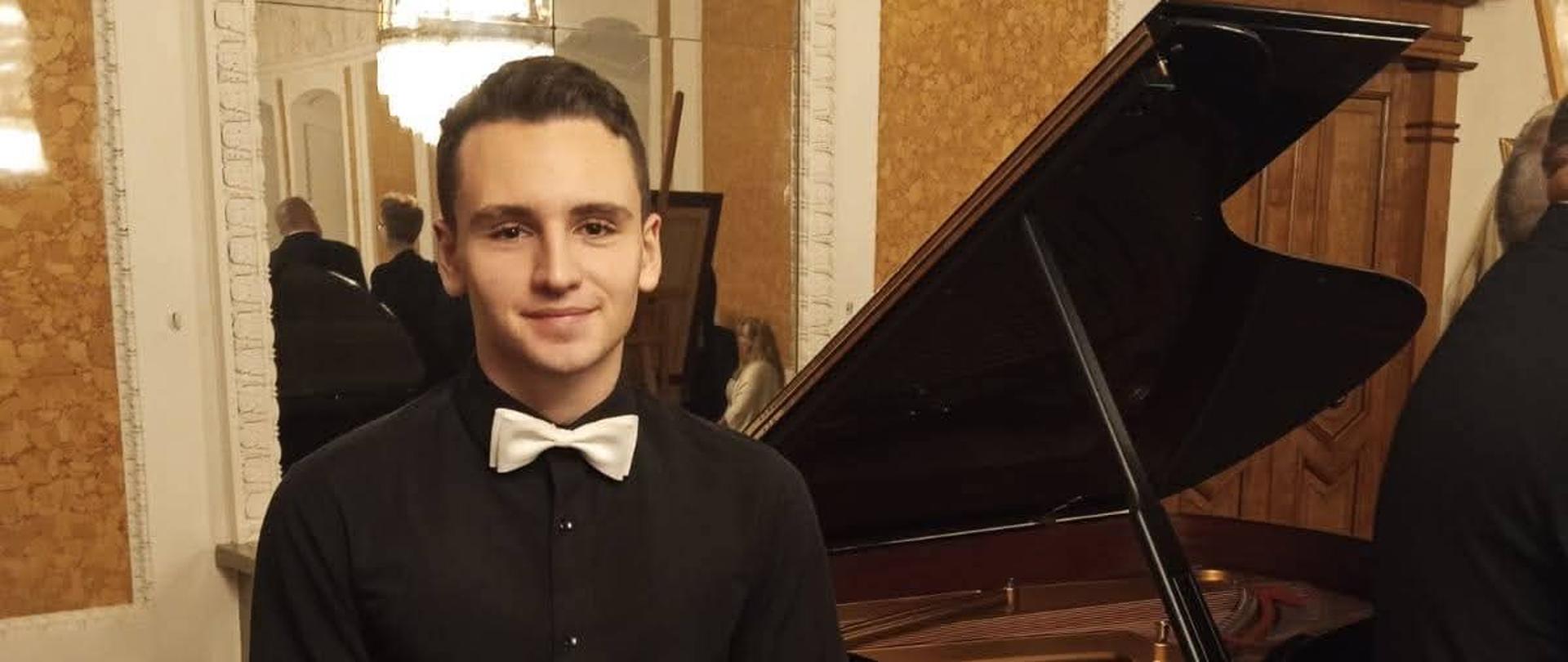 na zdjęciu na tle zabytkowych luster w białych ramach uczeń szkoły w czarnym ubraniu z białą muszka