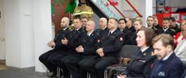 Strażacy w mundurach wyjściowych siedzą na krzesełkach w pomieszczeniu.