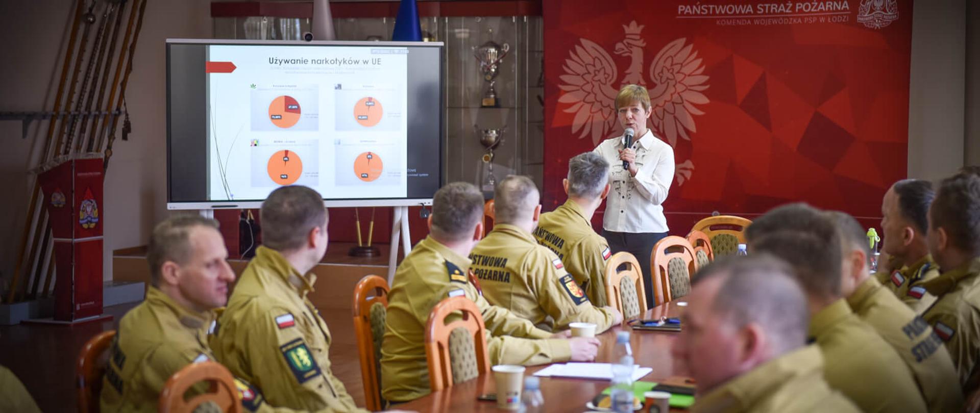kilku strażaków siedzących przy podłużnym stole, dalej stoi kobieta i omawia slajd wyświetlany na ekranie