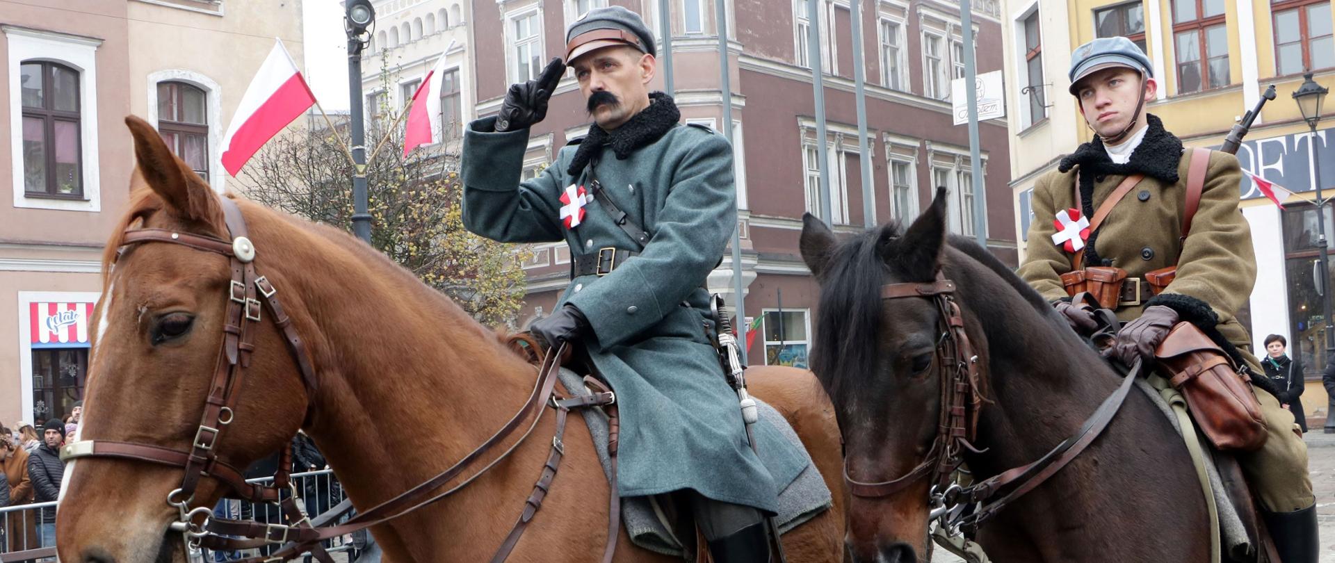 Zdjęcie przedstawia plac rynku miejskiego, na którym dwóch mężczyzn ubranych w historyczne mundury żołnierskie siedzi na koniach. W tle kamienice i zawieszone biało – czerwone flagi.