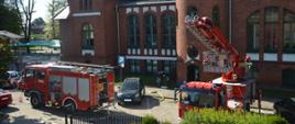 Ćwiczenia w Domu Kultury w Rawiczu. Ulica przed budynkiem, na niej dwa pojazdy pożarnicze biorące udział w ćwiczeniach. Na parkingu zaparkowane są samochody osobowe. W tle budynek. 