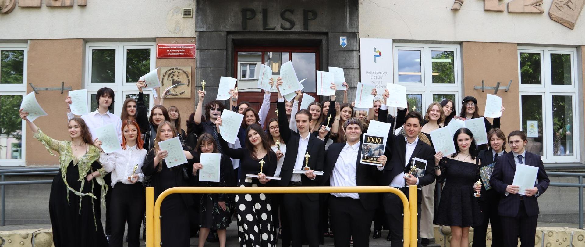 Zdjęcie: Na tle budynku szkoły uczennice i uczniowie w strojach galowych ze świadectwami w wyciągniętych dłoniach