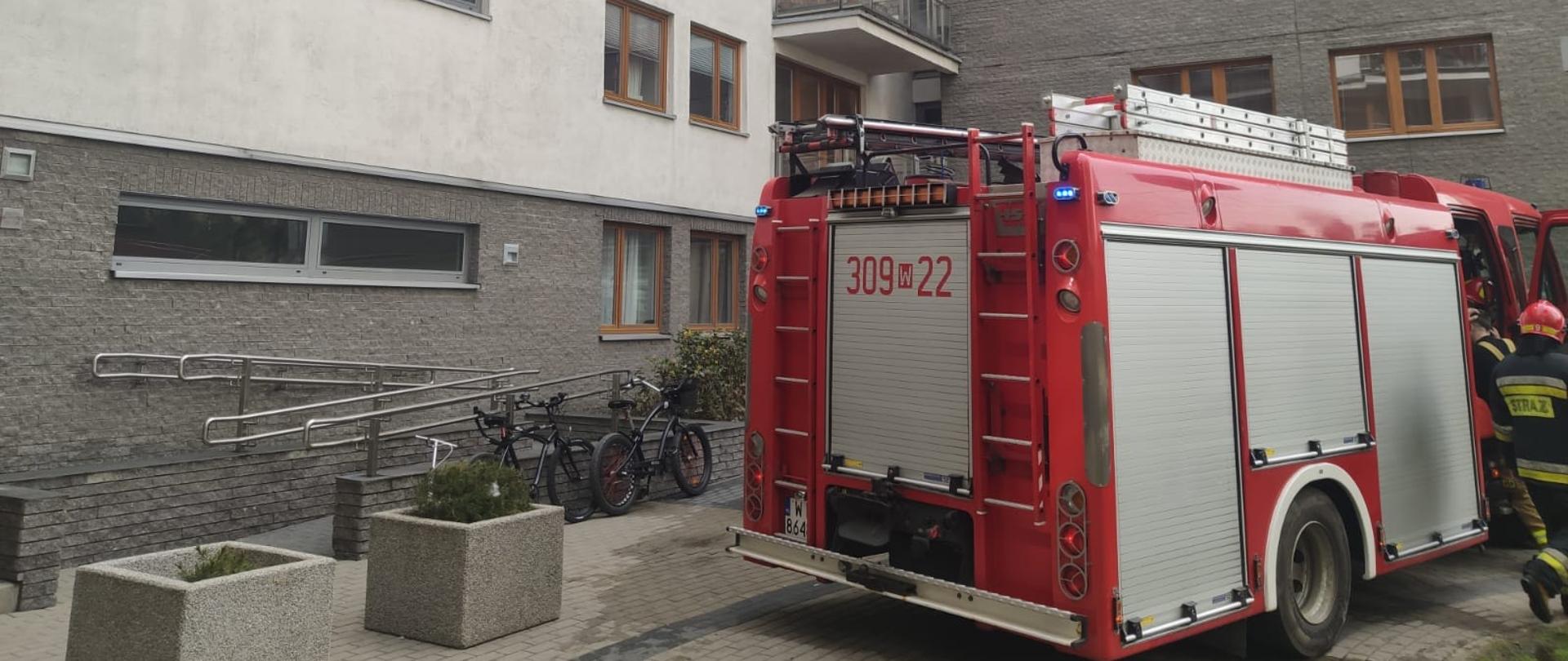 Na zdjęciu widoczny jest samochód pożarniczy, w tle widoczny jest budynek mieszkalny w którym miał miejsce pożar.