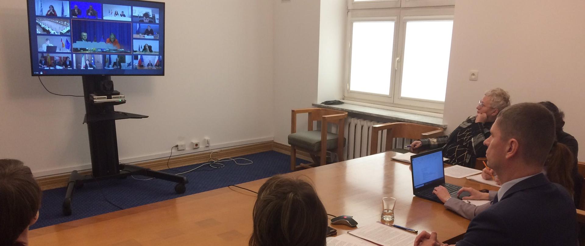 Minister edukacji siedzi przy stole i spogląda na duży monitor, gdzie widoczni są ministrowie edukacji państw Unii Europejskiej. 