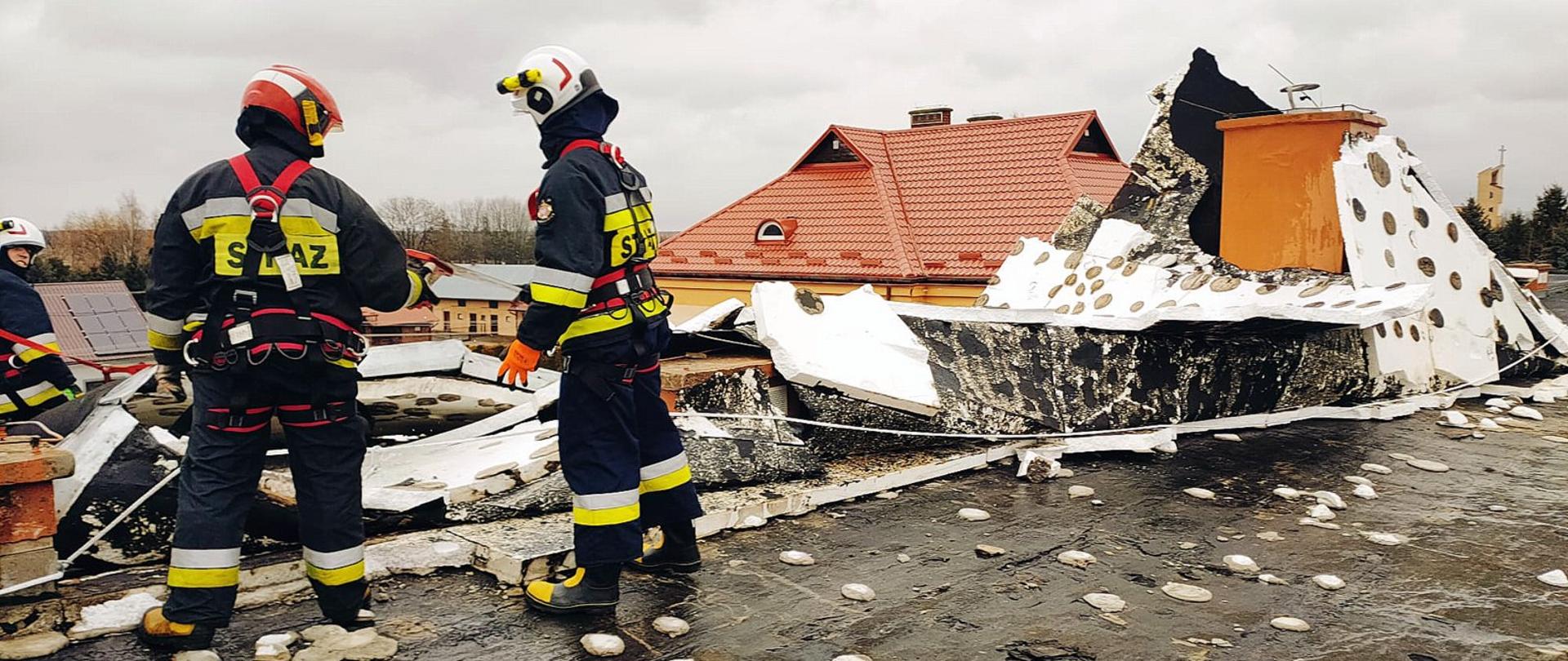 Zdjęcie zrobione na zewnątrz. Na uszkodzonym dachu stoi dwóch strażaków, którzy zabezpieczają uszkodzoną konstrukcję. Strażacy ubrani są ubrania specjalne, szelki oraz są zabezpieczeni linkami asekuracyjnymi. W tle widać zniszczone pokrycie dachowe. 