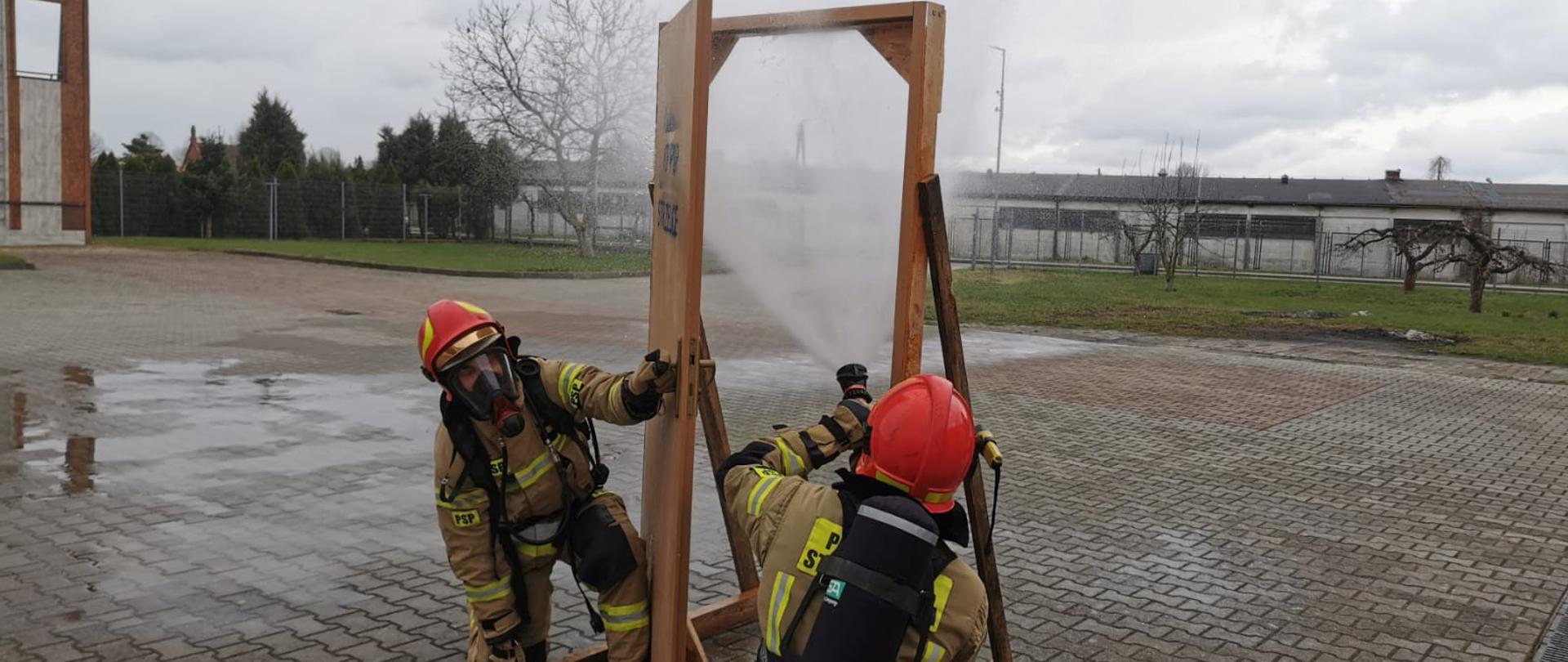 Fotografia przedstawia ujęcie placu z kostki polbrukowej. Na pierwszym planie widać dwóch strażaków w ubraniach specjalnych, hełmach, z założonymi aparatami ochrony układu oddechowego. Strażacy kucają przy atrapie drzwi do pomieszczenia. Jeden strażak kuca tyłem do fotografującego, kieruje wylot trzymanej w rękach prądownicy w górną część otworu drzwiowego, podaje rozproszony prąd wody. Drugi strażak, kuca przodem do fotografującego, uchyla drzwi umożliwiając podanie prądu wody pierwszemu. W tle widoczne tereny zielone w obrębie Jednostki ratowniczo-Gaśniczej PSP w Strzelcach Krajeńskich. Jest dzień, niebo jest zachmurzone.