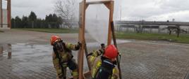 Fotografia przedstawia ujęcie placu z kostki polbrukowej. Na pierwszym planie widać dwóch strażaków w ubraniach specjalnych, hełmach, z założonymi aparatami ochrony układu oddechowego. Strażacy kucają przy atrapie drzwi do pomieszczenia. Jeden strażak kuca tyłem do fotografującego, kieruje wylot trzymanej w rękach prądownicy w górną część otworu drzwiowego, podaje rozproszony prąd wody. Drugi strażak, kuca przodem do fotografującego, uchyla drzwi umożliwiając podanie prądu wody pierwszemu. W tle widoczne tereny zielone w obrębie Jednostki ratowniczo-Gaśniczej PSP w Strzelcach Krajeńskich. Jest dzień, niebo jest zachmurzone.