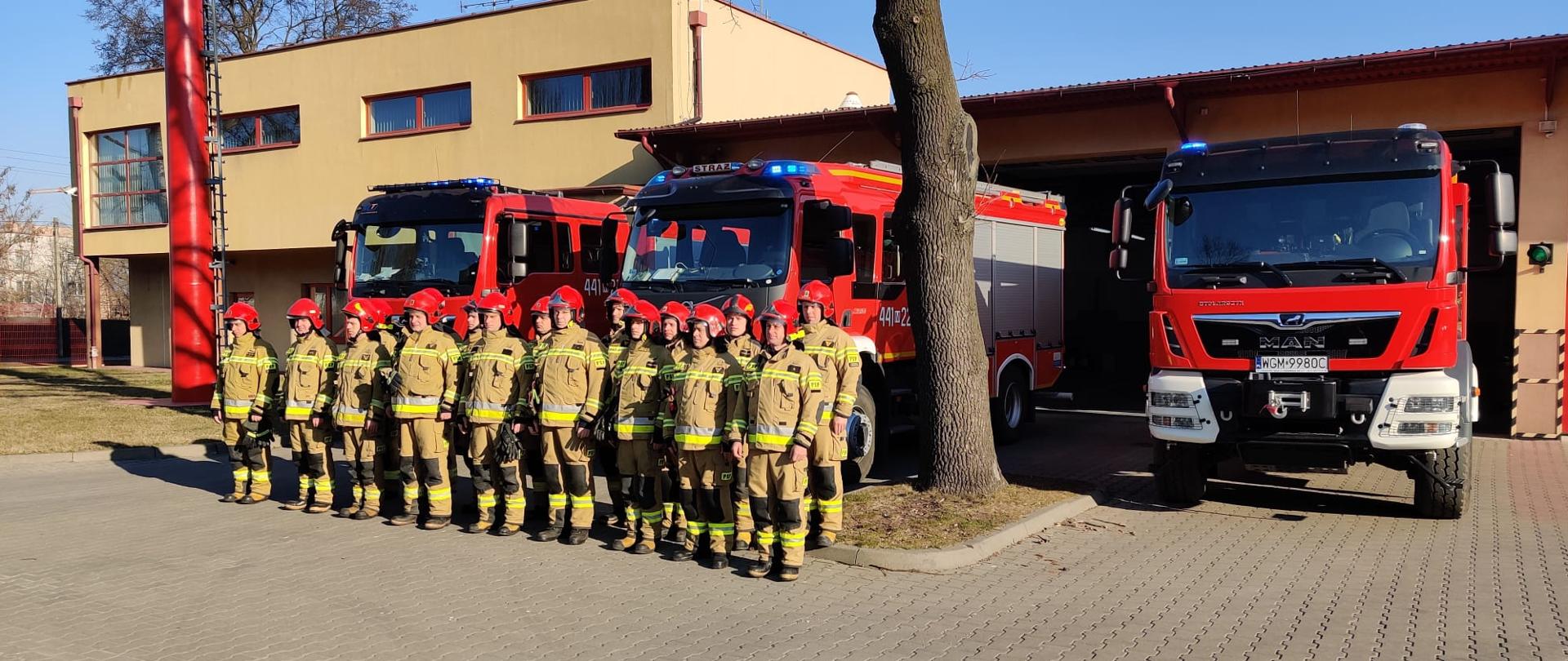 Strażacy PSP w Grodzisku Mzowieckim oddają hołd poległym strażakom Ukraińskim minutą ciszy. W tle samochody ratowniczo-gaśnicze z włączonymi tylko sygnałami świetlnymi oraz budynek KP PSP w Grodzisku Mazowieckim. 