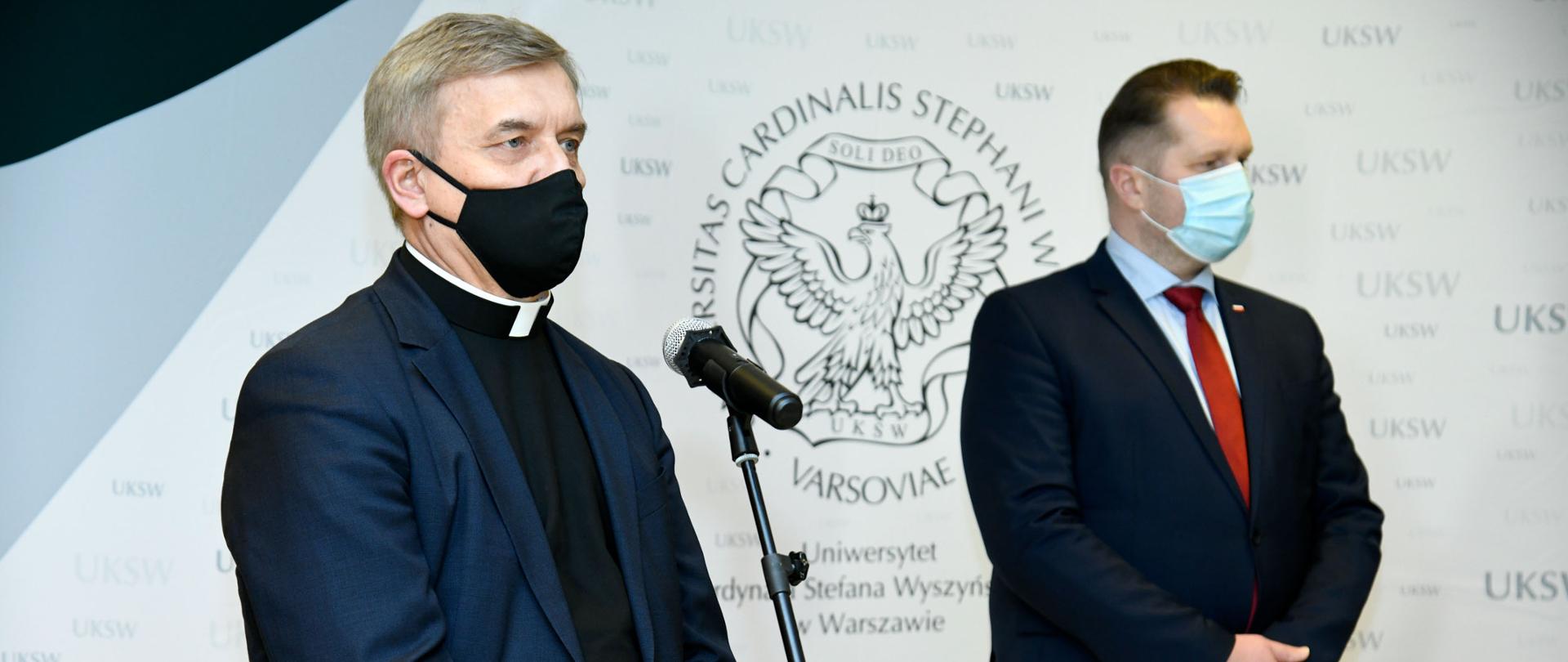 Na pierwszym planie rektor Uniwersytetu Kardynała Stefana Wyszyńskiego w Warszawie, który stoi przy mikrofonie. Na twarzy ma czarną maseczkę. Obok minister Przemysław Czarnek w maseczce na twarzy. Obaj ubrani w garnitur. W tle baner UKSW. 