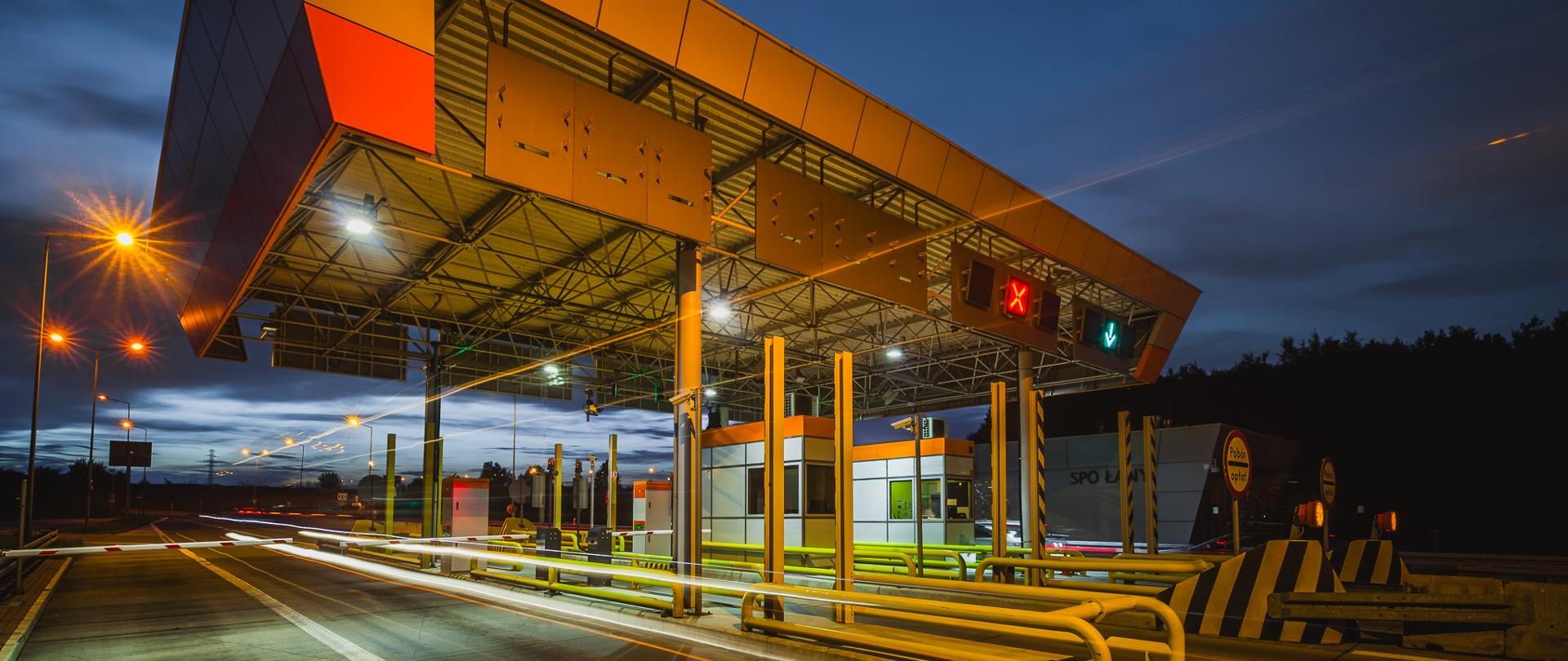 Miejsce Poboru Opłat na autostradzie A4 w porze wieczornej, z charakterystycznym zadaszeniem nad pasami ruchu, sygnalizacją świetlną nad bramkami oraz śladami świateł przejeżdżających pojazdów na tle ciemniejącego nieba.