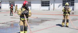 Na pierwszym planie dwóch strażaków podczas egzaminu praktycznego, w umundurowaniu specjalnym, z podpięta butlą na plecach oraz hełmie, zwijający wąż. W tle strażak zwijający wąż