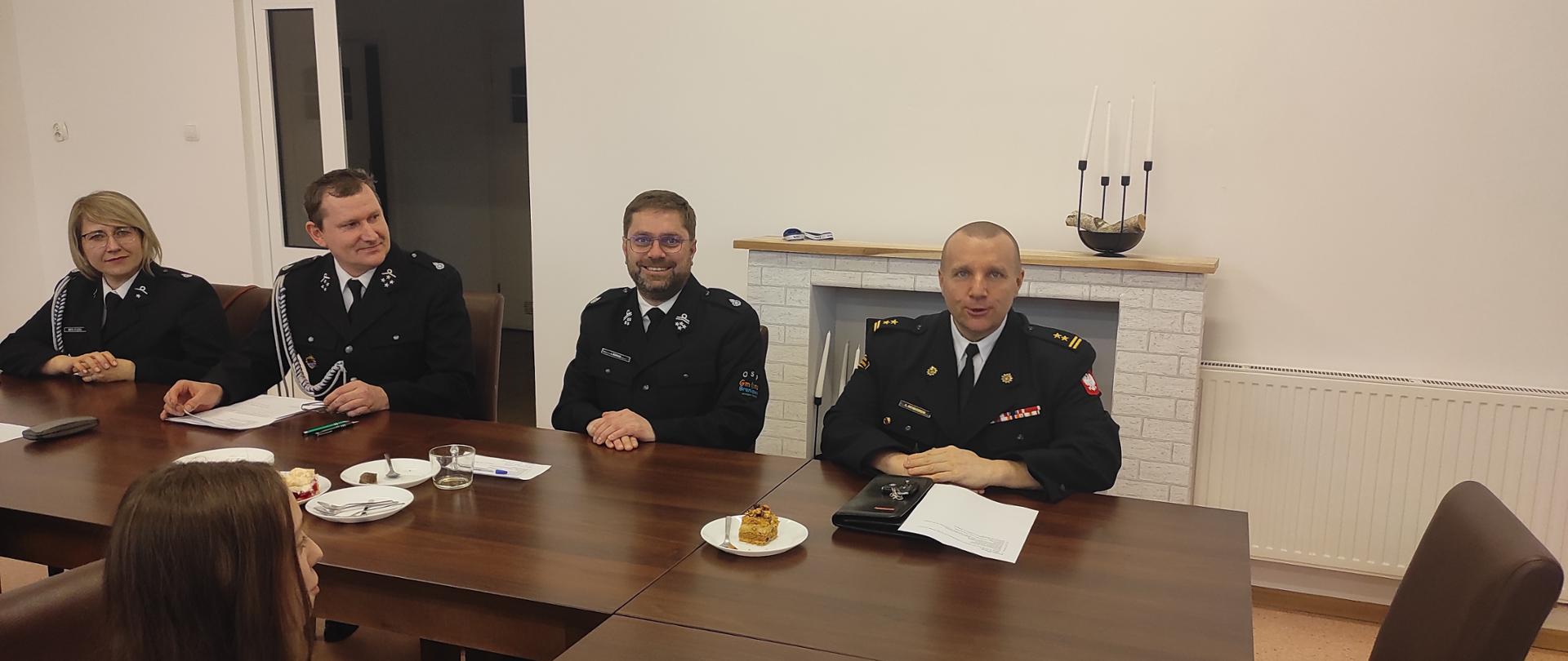 Zdjęcie zrobione na świetlicy. Przy brązowym stole, na tle białej ściany siedzi czterech strażaków w mundurach wyjściowych. Uśmiechają się do zdjęcia.