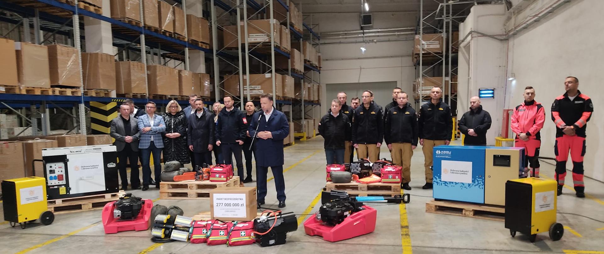 Grupa osób, w tym strażacy i przedstawiciele administracji, stoi w magazynie przemysłowym przy paletach z nowym sprzętem ratowniczym; widoczne są zestawy medyczne, dmuchawy, agregaty i pomarańczowe pilarki.