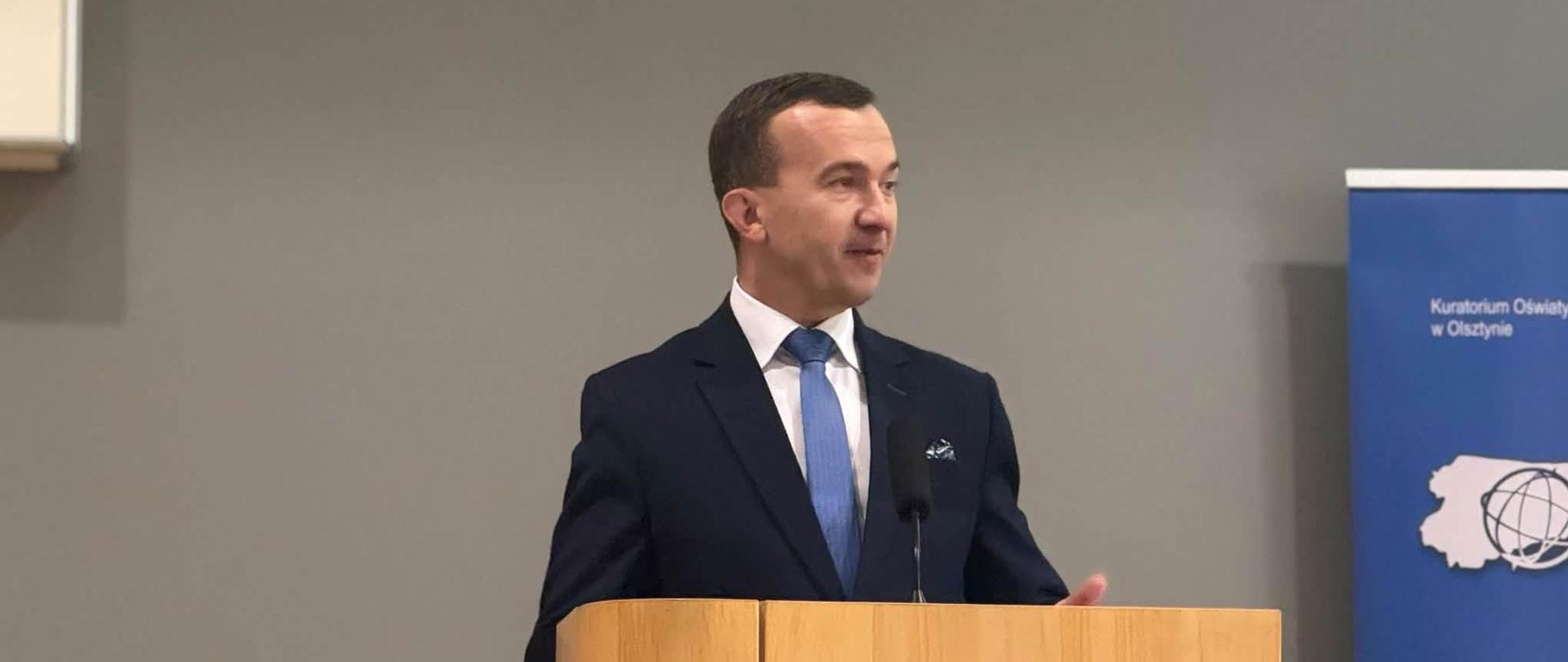 Mężczyzna w granatowym garniturze stoi przy mównicy na scenie 