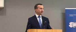 Mężczyzna w granatowym garniturze stoi przy mównicy na scenie 