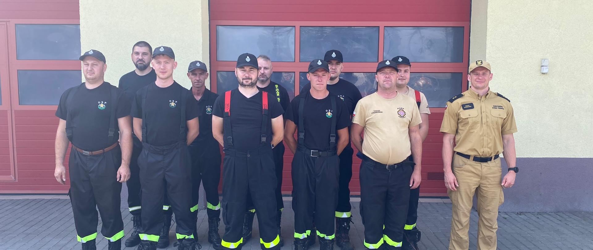 przed garażem stoi 11 druhów OSP w ubraniach koszarowych, po prawej stronie stoi umundurowany funkcjonariusz PSP