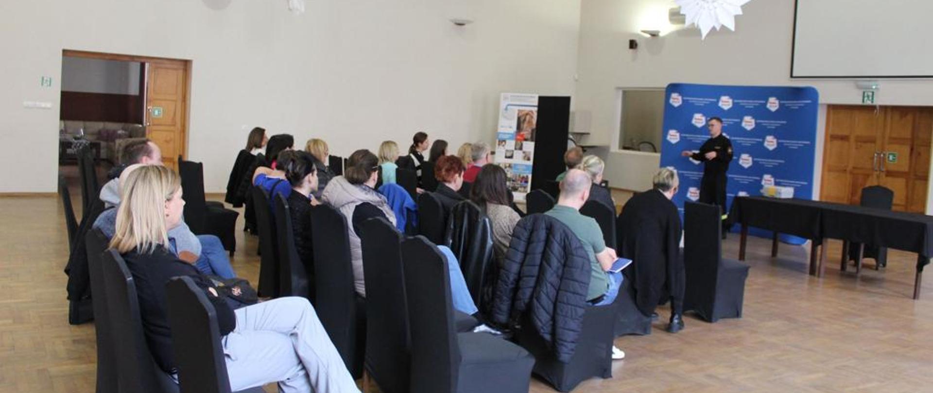 Na oświetlonej sali siedzi grupa osób, przed nimi stoi mężczyzna w mundurze straży pożarnej przemawiający do zabranych, za strażakiem stoi niebieski baner CKiW OHP w Pleszewie