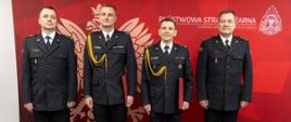 Wspólne zdjęcie grupowe oficerów Państwowej Straży Pożarnej na tle czerwonego banneru Państwowej Straży Pożarnej z białym Godłem RP oraz napisem Państwowa Straż Pożarna, logiem PSP oraz napisem Komenda Wojewódzka PSP w Opolu. Funkcjonariusze w mundurach wyjściowych.
Stoją od lewej: st. bryg. Arkadiusz Kuśmierski, mł. bryg. Szymon Jędrzejas, st. bryg. Piotr Dudek oraz st. bryg. Paweł Kielar.