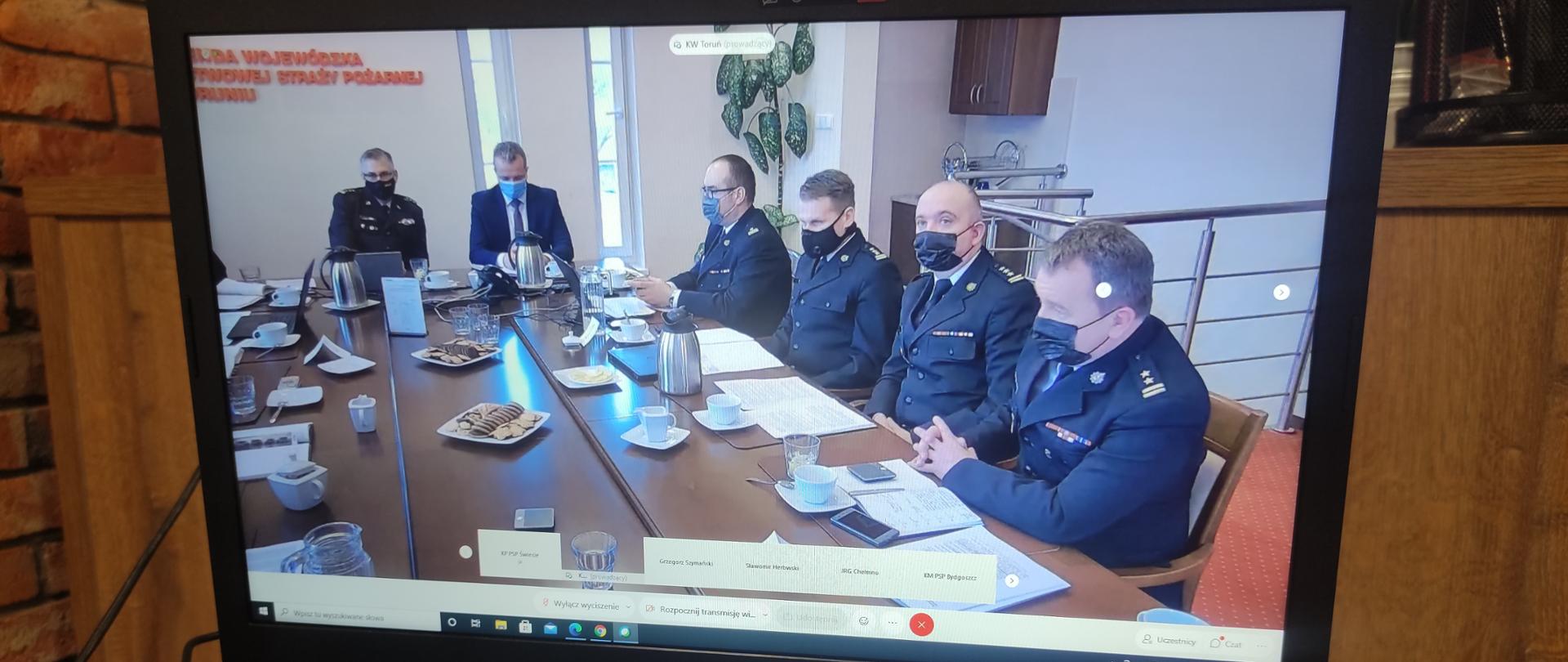 Na zdjęciu laptop w którym widać siedzących przy stole funkcjonariuszy Komendy Wojewódzkiej Państwowej Straży Pożarnej w Toruniu oraz Wojewodę Kujawsko-Pomorskiego Pana Mikołaja Bogdanowicza